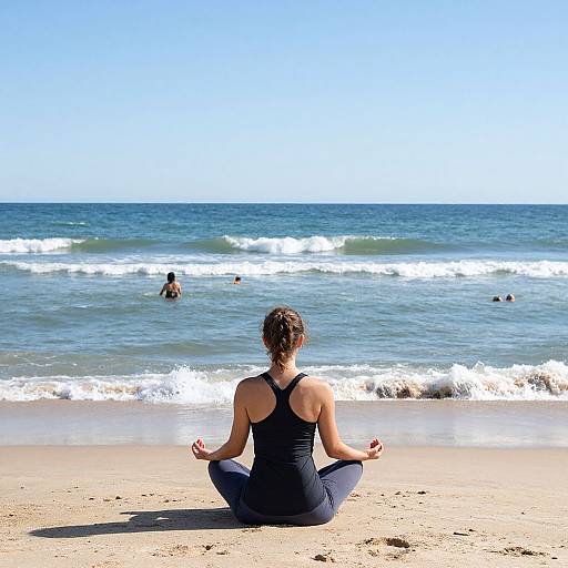 Photograph of a woman with brown hair in a black tank top and gray leggings, sitting cross-legged on a sunny beach, meditating with her back