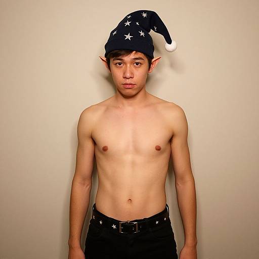 Photograph of a young, shirtless Asian man with pointed elf ears, wearing a black starry hat and black pants, standing against a plain white