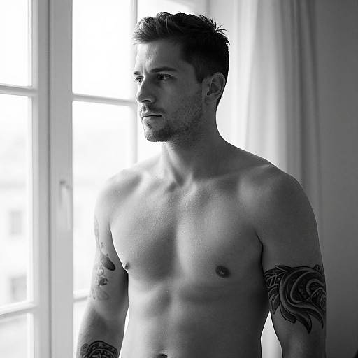 Photograph of a shirtless, muscular, tattooed man with short dark hair, standing in a brightly lit room with a window behind him. Mon