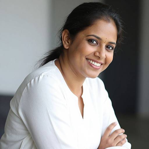 Photograph of a smiling Indian woman with dark hair in a ponytail, wearing a white blouse, standing indoors with blurred background.