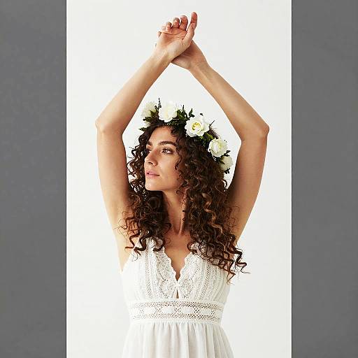 Photograph of a curly-haired woman with a white flower crown, wearing a white lace dress, arms raised, against a plain white background.