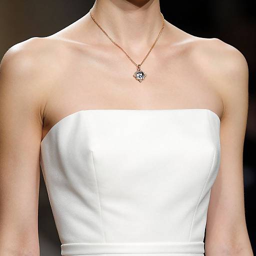 Photograph of a fair-skinned woman with a slender neck, wearing a strapless white dress and a delicate gold necklace with a pendant, against a