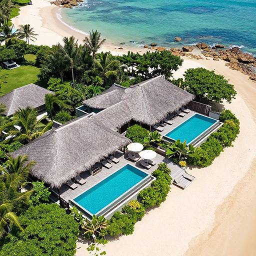 Aerial photograph of a tropical beach resort with two blue pools, thatched-roof villas, lush greenery, and a sandy beach with clear