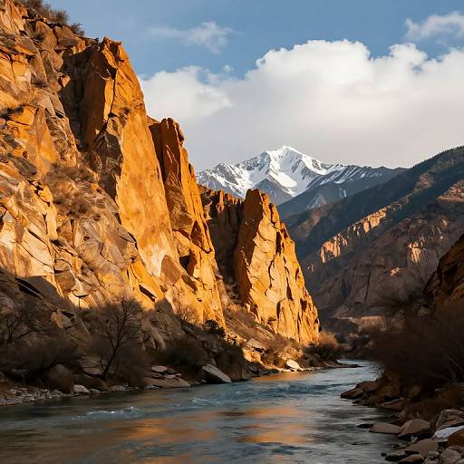Photograph of a sunlit canyon with towering orange rock cliffs, a flowing river, and snow-capped mountains in the background.