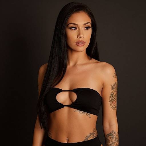 Photograph of a tattooed, light-brown-skinned woman with long black hair, wearing a black, cut-out strapless top, standing against