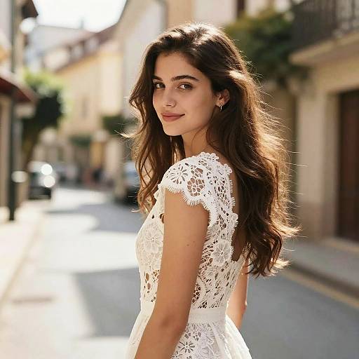 Photograph of a young woman with long, wavy brown hair, wearing a white lace dress, standing on a sunlit, narrow European street.