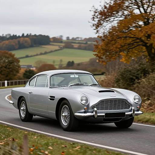 Photograph of a silver classic Aston Martin DB5 driving on a curving country road with autumn foliage in the background.