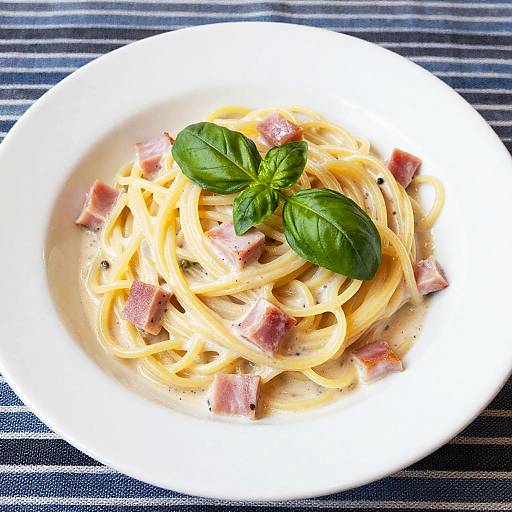 Photograph of creamy fettuccine pasta with pink ham chunks and fresh basil leaves on a white plate, set on a blue-striped tablecloth.
