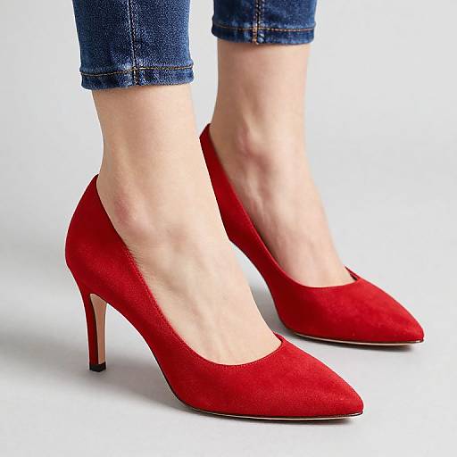 Photograph of a woman's feet wearing red suede pointed-toe high heels with slim black heels, paired with cuffed blue denim jeans, against a