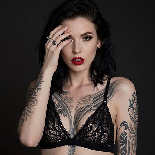Photograph of a pale-skinned woman with black hair, red lipstick, black lace bra, and extensive tattoos, touching her forehead with a hand adorned
