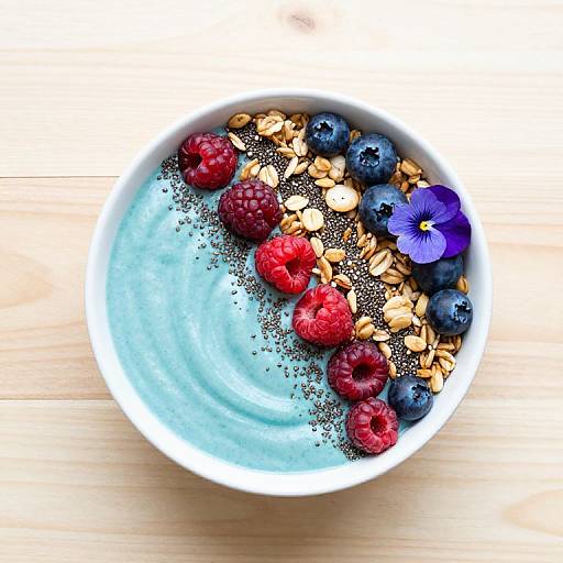 Top-down photo of a white bowl with blue yogurt, black chia seeds, red raspberries, blueberries, almonds, and a purple flower on a