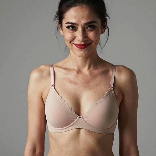 Photograph of a smiling woman with light skin, dark hair in a bun, wearing a light beige lace-trimmed bra, against a plain gray