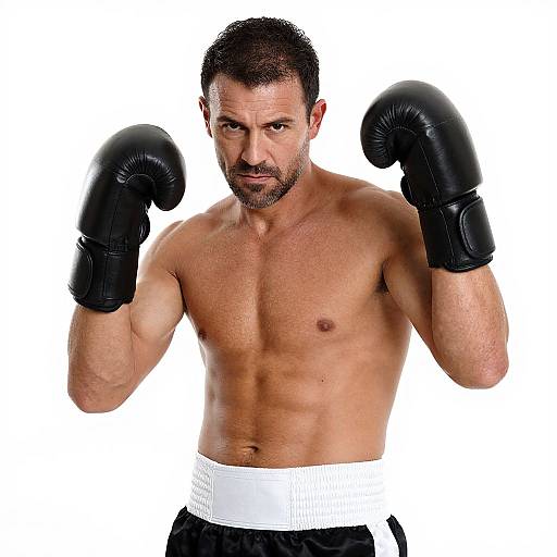 Photograph of a muscular, tan-skinned, dark-haired man with a beard, wearing black boxing gloves and black shorts with a white waistband,