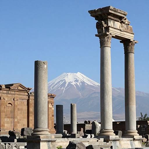 Photograph of ancient Greek-style columns with ornate capitals in front of a snow-capped mountain, under a clear blue sky.