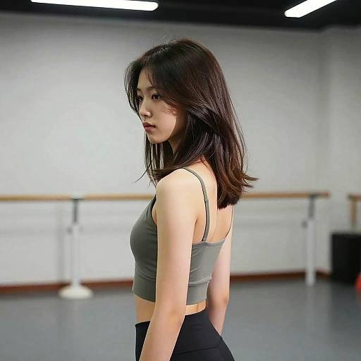 Photograph of an Asian woman with shoulder-length black hair, wearing a gray crop top and black pants, standing in a modern dance studio with white walls