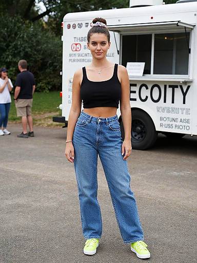 Photograph of a young woman with a bun, wearing a black crop top, high-waisted blue jeans, and neon yellow sneakers, standing in