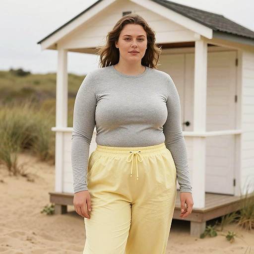 Photograph of a curvy woman with light skin, brown hair, gray long-sleeve top, and yellow pants, standing in front of a