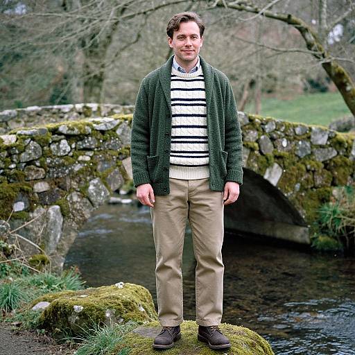 Photograph of a man with short brown hair, wearing a green cardigan, white striped shirt, beige pants, and brown shoes, standing on a