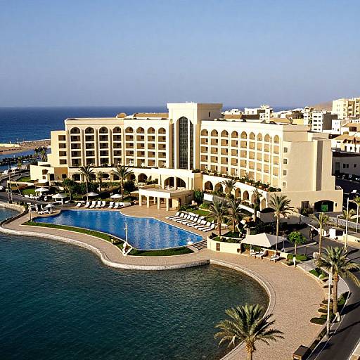 Aerial photograph of a luxurious, cream-colored beachfront hotel with a curved pool, palm trees, sun loungers, and clear blue ocean.