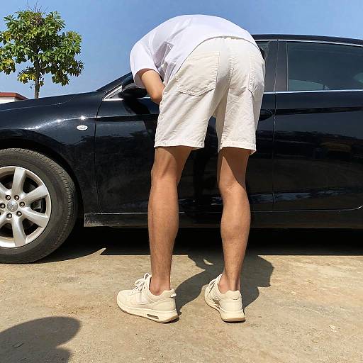 Photograph of a man in white shorts and sneakers, bending over to inspect a black car's front wheel on a sunny day.