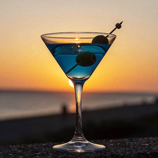 Photograph of a martini glass with blue liquid, a black olive, and a toothpick, silhouetted against a vibrant sunset.