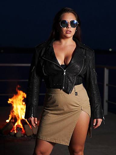 Nighttime photograph of a woman with dark hair, wearing round sunglasses, black leather jacket, and beige skirt with high slit, standing in front of a