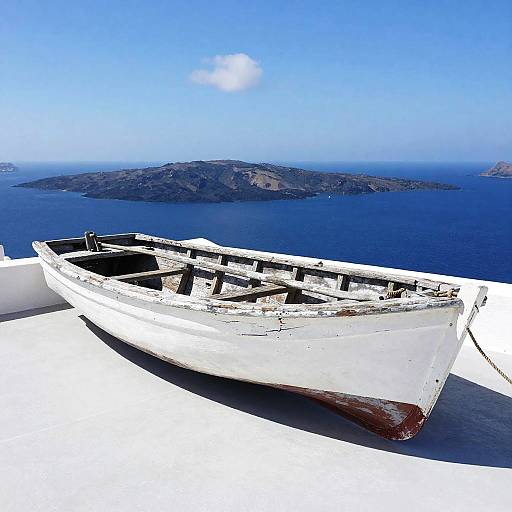 Photograph of a weathered white wooden boat with red bottom, anchored on a bright white terrace, overlooking a deep blue sea and distant mountainous island