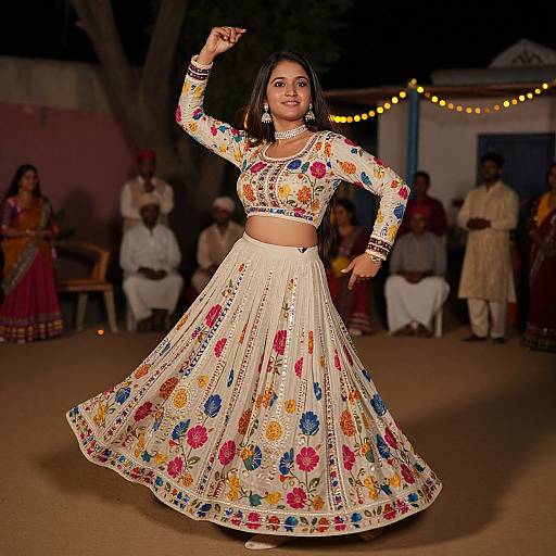 Photograph of a smiling Indian woman in a colorful, floral embroidered traditional outfit, mid-dance, on a lit runway, with audience in background.