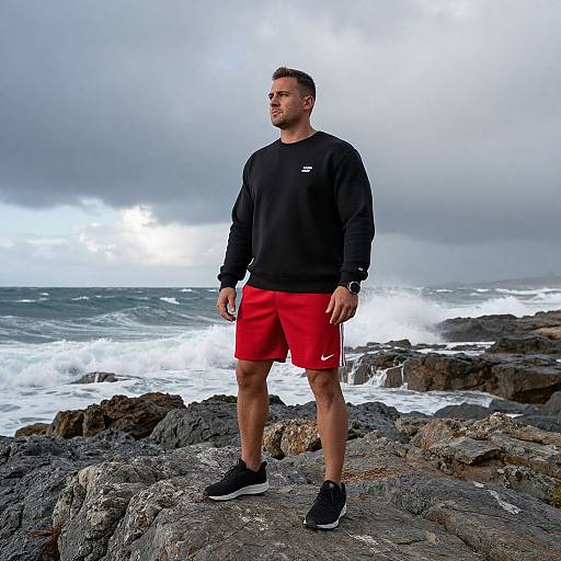 Photograph of a muscular man with short brown hair, wearing a black sweatshirt, red Nike shorts, and black sneakers, standing on rocky coastline with