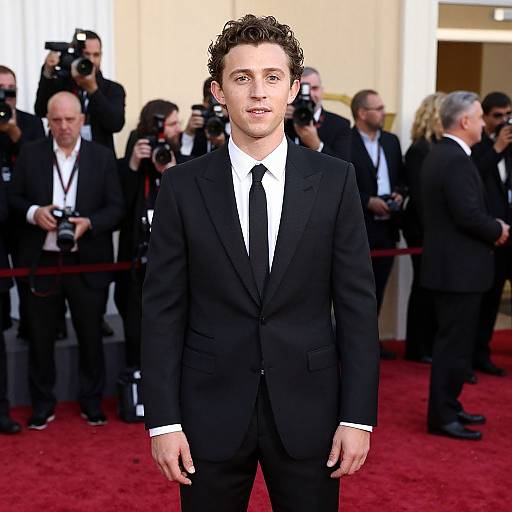 Photograph of a young, white male with curly brown hair, wearing a black suit, white shirt, and black tie, standing on a red carpet