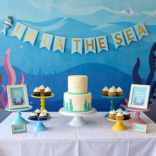 Under-the-sea themed birthday table with white cake, cupcakes on colorful stands, framed photos, 