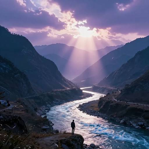 Photograph of a solitary figure standing on a rocky cliff, overlooking a sparkling, sunlit river winding through misty, purple-tinged mountains at