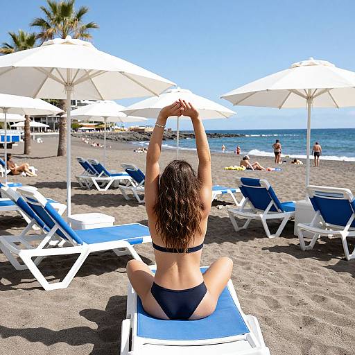 Photograph of a woman in a black bikini, sitting on a blue lounge chair, stretching with arms raised, on a sunny beach with white umbrellas