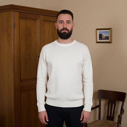 Photograph of a bearded man with short dark hair, wearing a white sweater and black pants, standing in a warmly lit room with wooden cabinets,