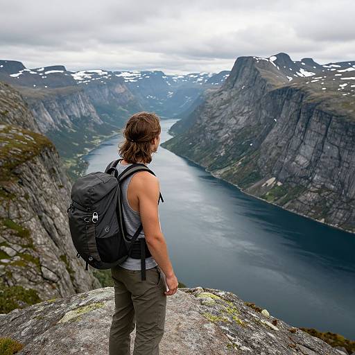 Photograph of a muscular man with shoulder-length brown hair, wearing a gray tank top and green pants, standing on a rocky outcrop, facing a