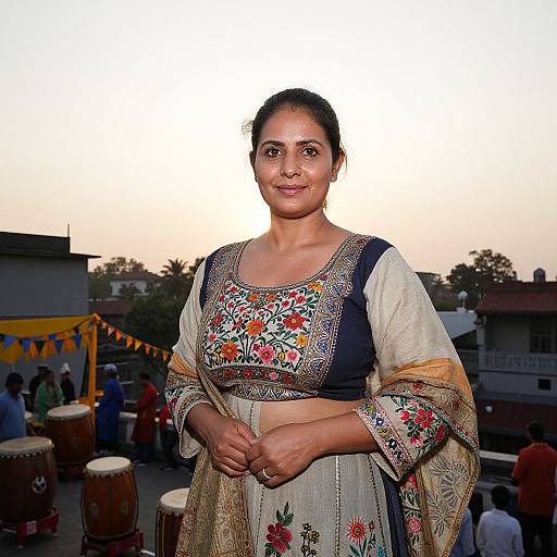 Photograph of a smiling Indian woman in a traditional floral embroidered saree, standing outdoors at dusk with a drumming group in the background.
