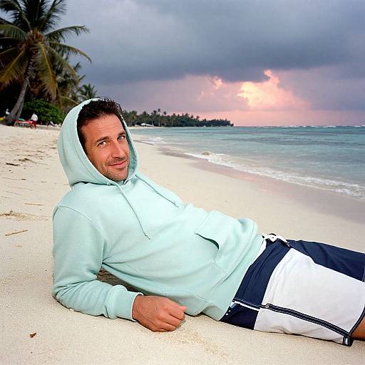 Photograph of a smiling man in a light blue hoodie and black-and-white shorts, lying on a sandy beach with palm trees and a cloudy, sunset