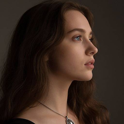 Photograph of a young woman with fair skin, blue eyes, and long dark brown hair, wearing a silver necklace, in profile against a dark background