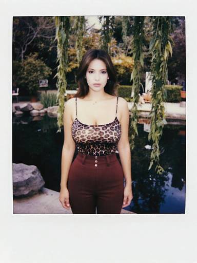 Photograph of a young woman with medium skin tone, dark wavy hair, wearing a leopard print top and high-waisted brown pants, standing