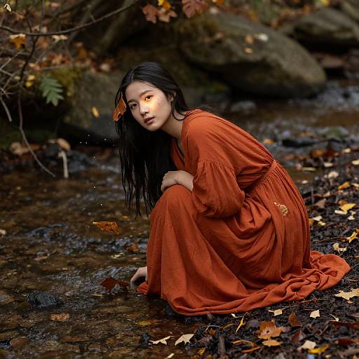 Photograph of an Asian woman with long black hair, wearing an orange dress, squatting by a forest stream, surrounded by autumn leaves and rocks,