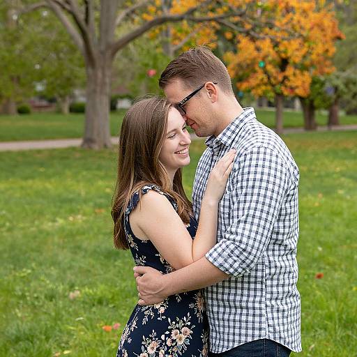 Photograph of a smiling couple embracing outdoors in a grassy park with autumn trees; woman in floral dress, man in checkered shirt.