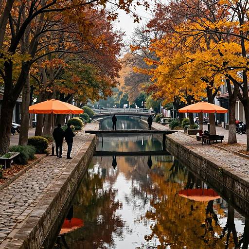 Photograph of a cobblestone canal lined with autumn trees, orange umbrellas, and reflections, with a bridge in the background.