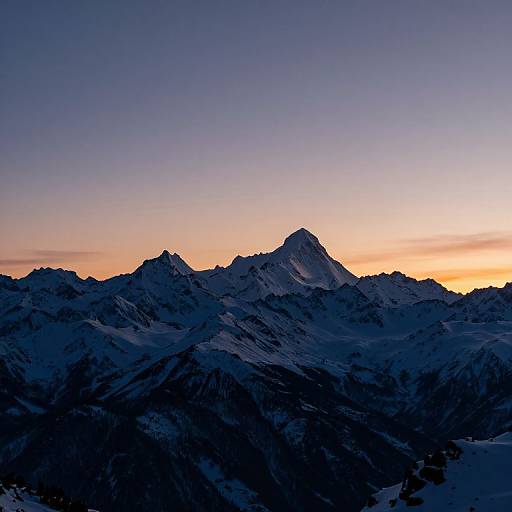 Photograph of a snow-capped mountain range at sunset, with dark blue mountains silhouetted against a gradient sky of orange, yellow, and