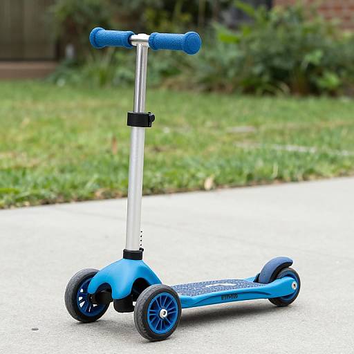 Photograph of a blue plastic scooter with black handle grips, silver stem, and black wheels on a concrete sidewalk. Green grass and blurred garden background.