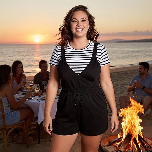Photograph of a smiling woman with curly brown hair, wearing a black pinafore over a striped shirt, standing in front of a sunset beach fire