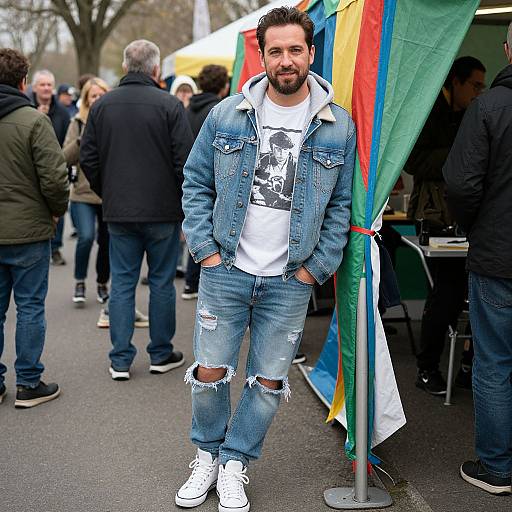 Photograph of a bearded man with dark hair, wearing a distressed blue denim jacket, white graphic tee, ripped jeans, and white sneakers, standing