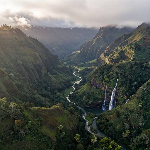 Aerial photograph of lush, green mountainous landscape with a winding river, twin waterfalls, and sunlight piercing through clouds.