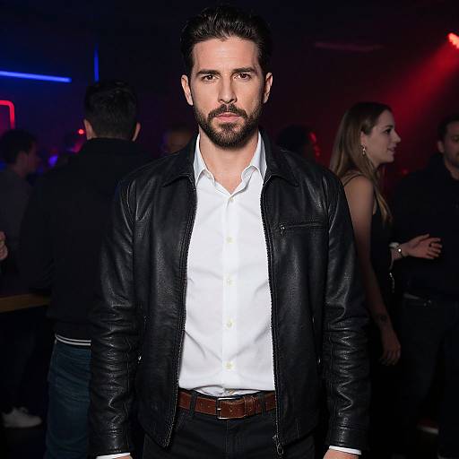 Photograph of a bearded man with dark hair, wearing a black leather jacket over a white shirt, standing in a dimly lit club with colorful