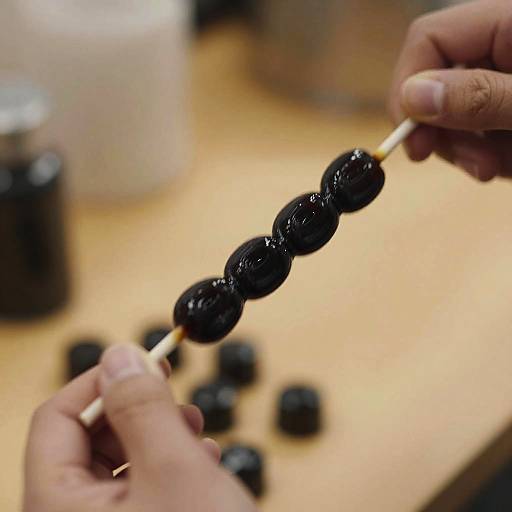 Photograph of hands holding a skewer with glossy black grilled round meat pieces, against a blurred wooden surface with scattered black round objects.
