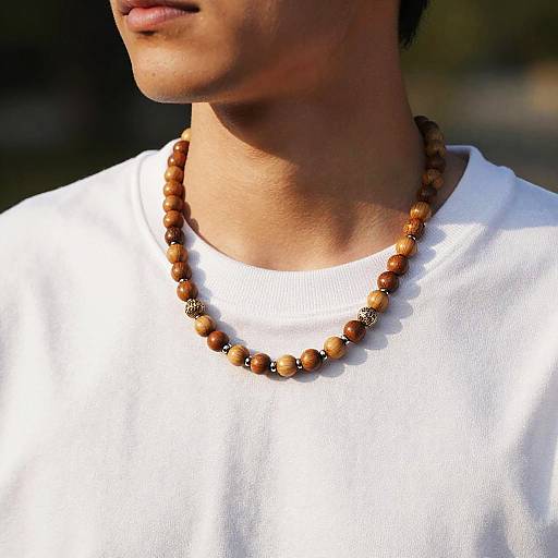 Photograph of a man's lower face and neck, wearing a white shirt and a wooden bead necklace with a black decorative bead. Sunlight highlights his